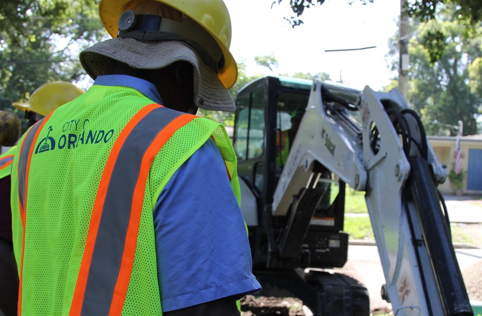 A photo of a City employee wearing a hi-visibility vest look at a white excavator